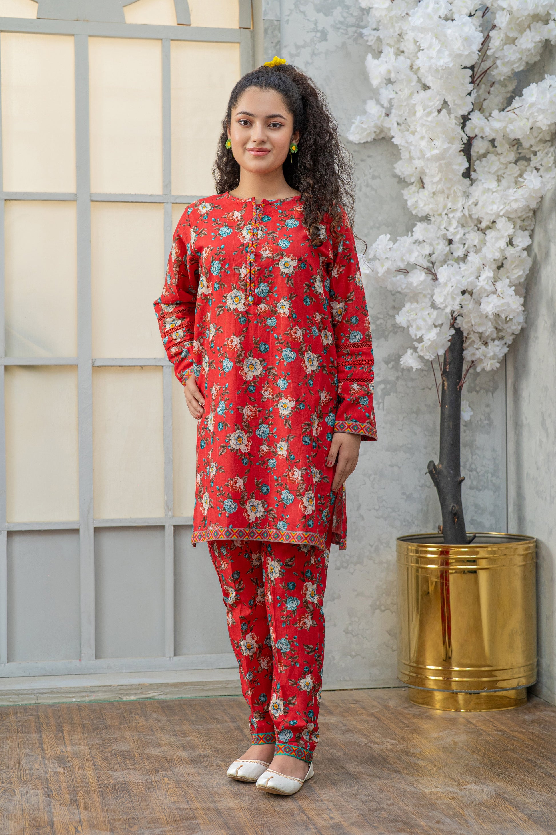 Woman wearing a red floral outfit standing in front of a decorative plant and window.
